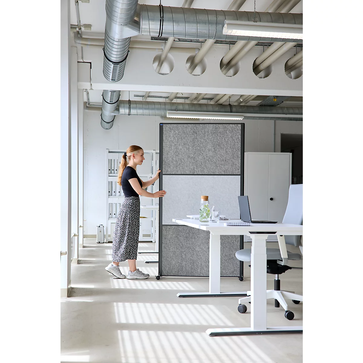 Eine junge Frau positioniert einen grauen, dreigeteilten Raumtrenner in einem modernen Büro mit Industrie-Stil.