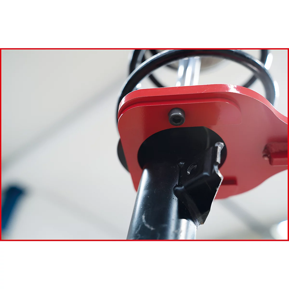 Nahaufnahme eines roten, metallischen Werkzeugs, das an einem Stoßdämpfer befestigt ist. Im Hintergrund ist eine Feder und ein unscharfer heller Raum zu sehen.