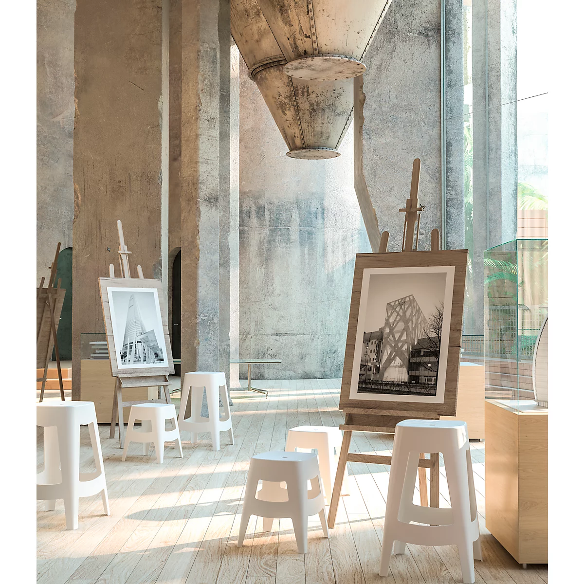Ein Atelier mit Staffeleien, Bildern und Hockern. Im Hintergrund ein großes metallenes Element.
