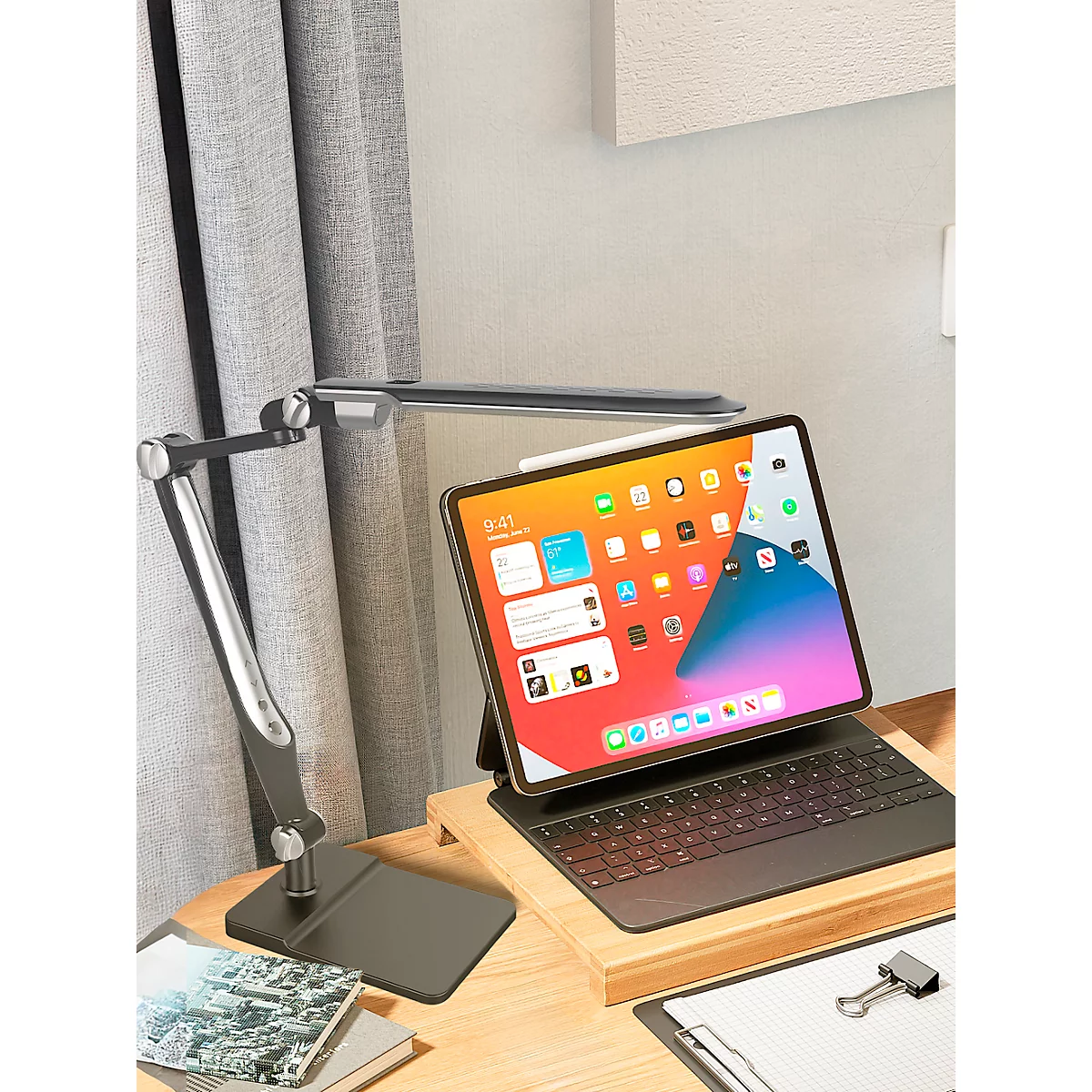 Ein iPad mit Tastatur und Stift steht auf einem Holzsockel. Eine Lampe leuchtet das Tablet aus, es liegt auf einem Holzschreibtisch.
