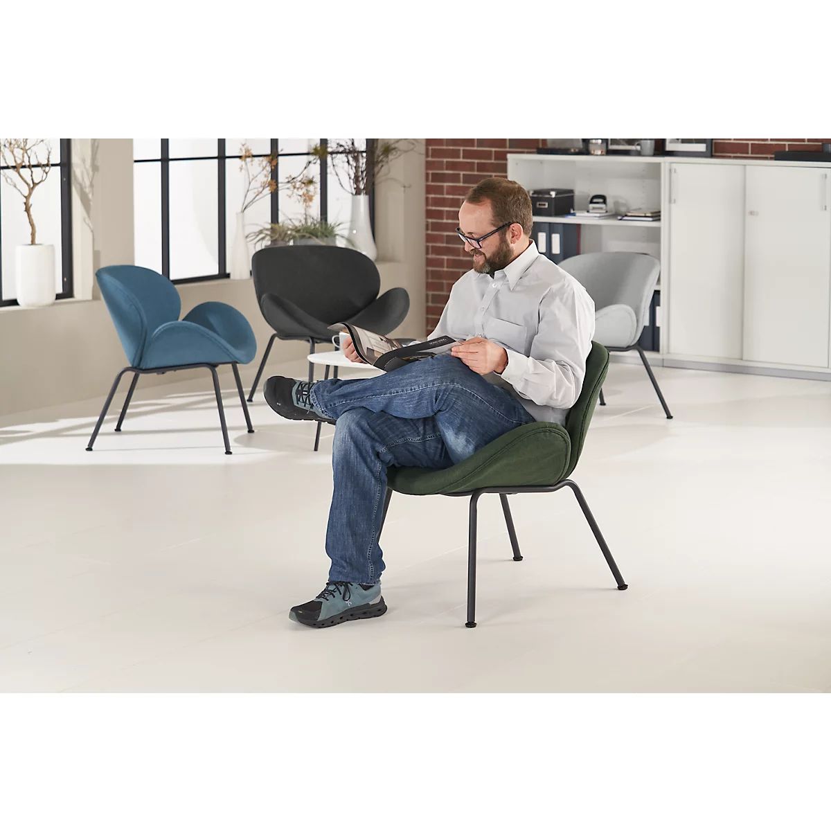 Ein Mann in Jeans und Hemd sitzt in einem grünen Sessel und liest. Im Hintergrund weitere Stühle und ein Fenster.