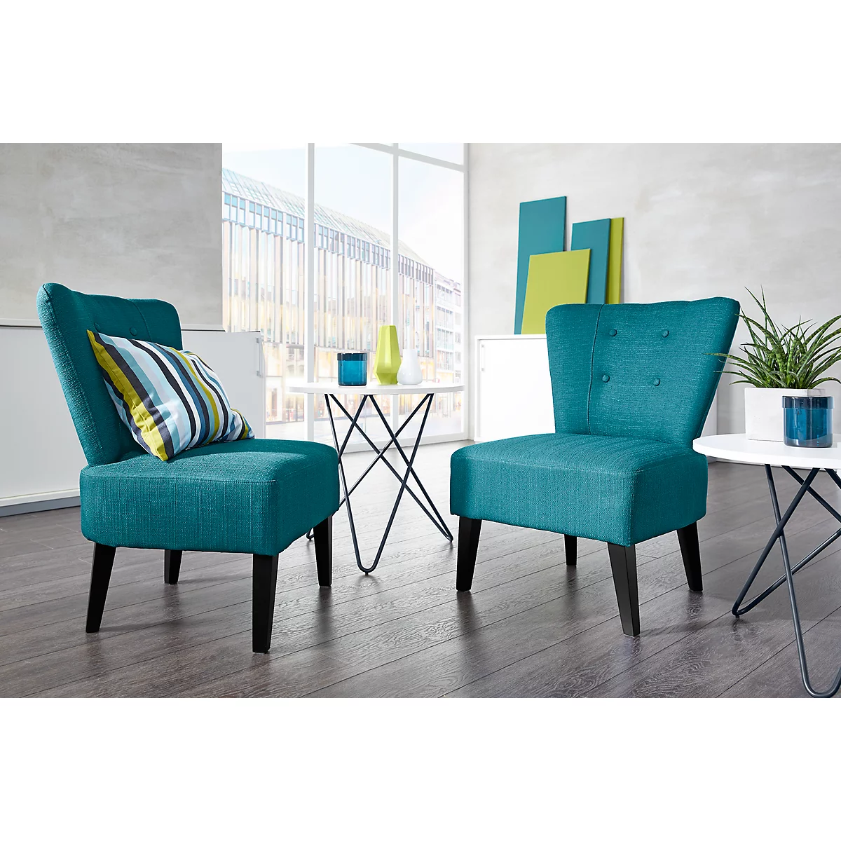 Dos sillones turquesa con patas de madera en un salón luminoso. Un cojín descansa sobre un sillón. En el fondo: ventana, plantas, decoración.