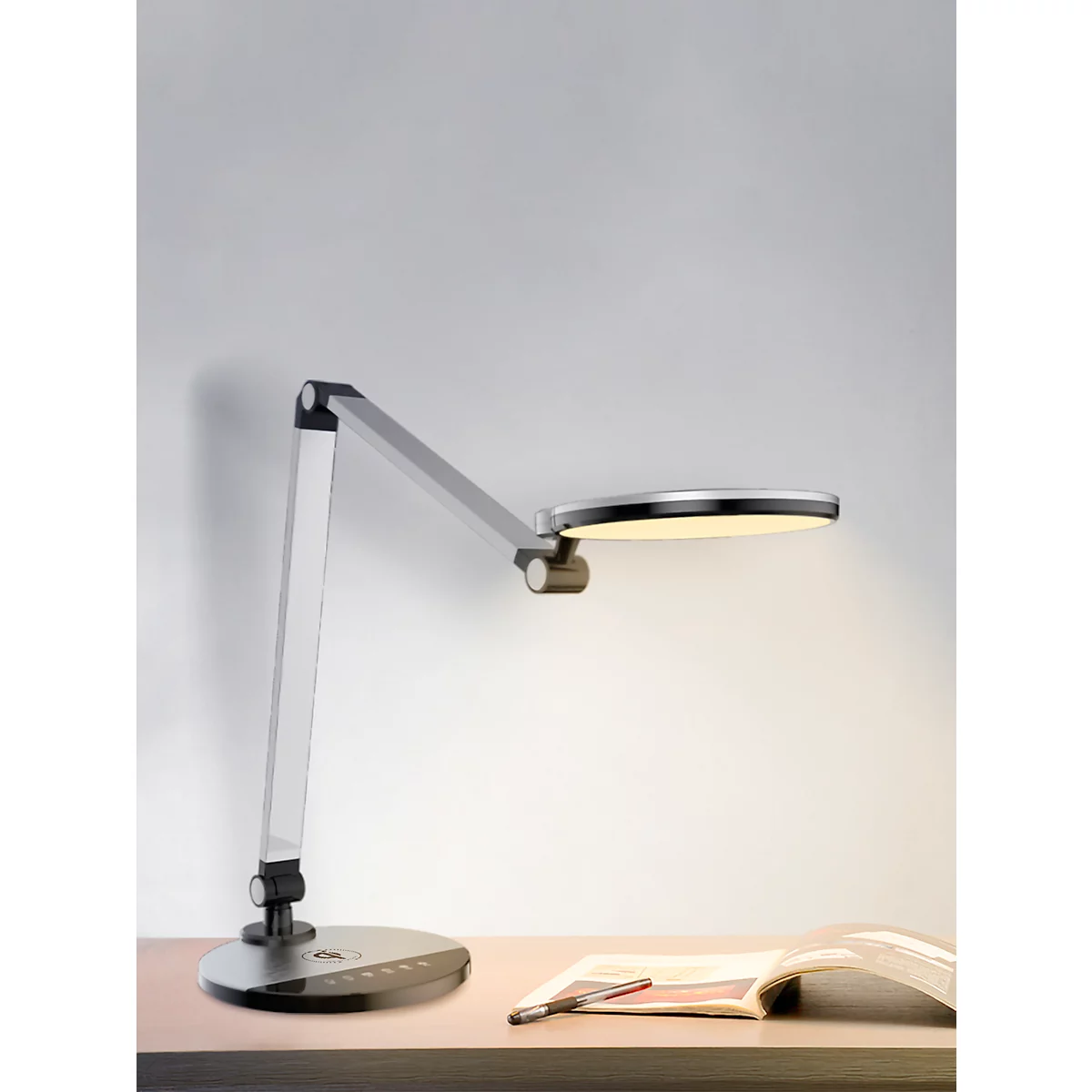Een zilveren bureaulamp met een ronde lampkop die een gelige verlichting geeft. Op een bureau naast een open boek.