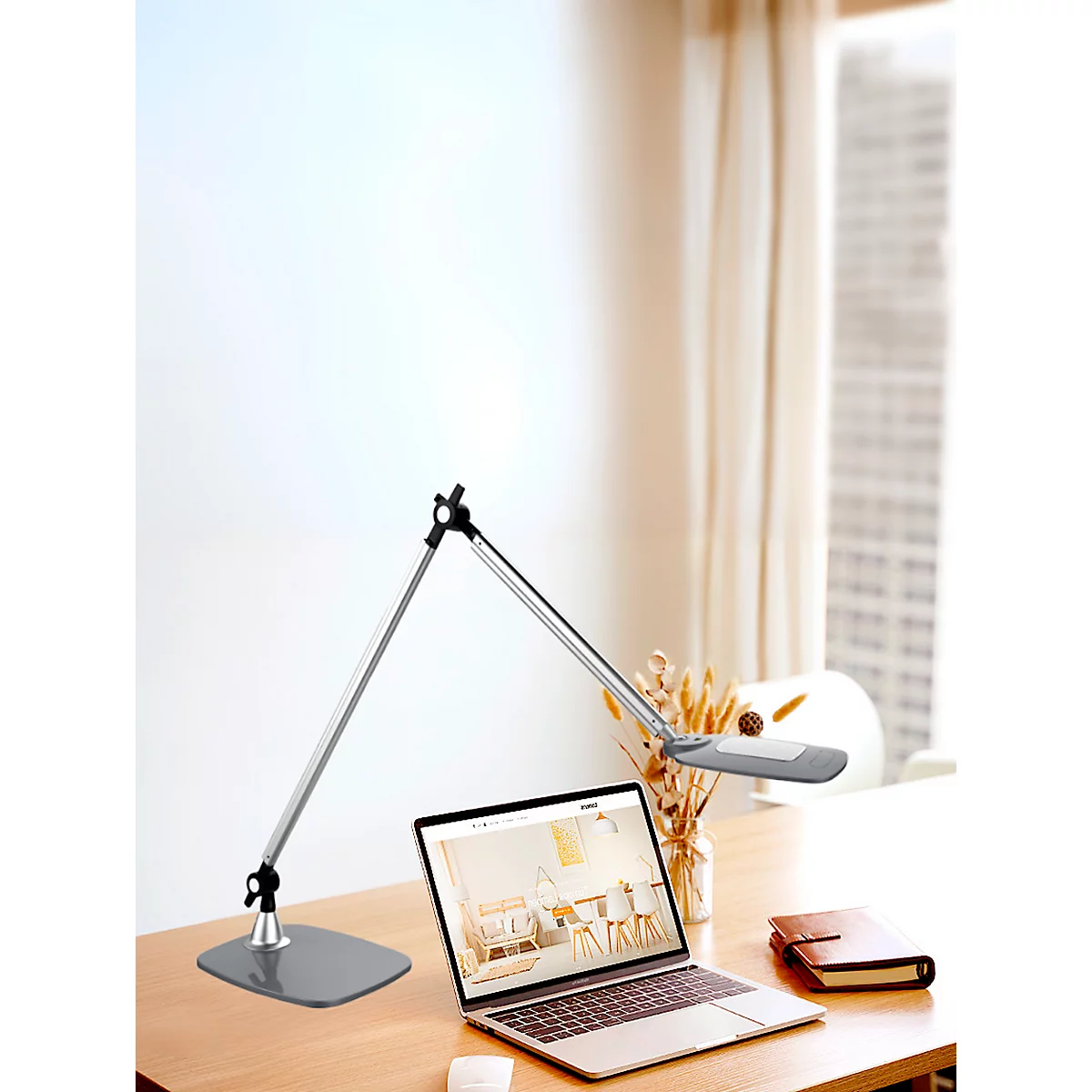 Zilveren bureaulamp op tafel naast laptop, notitieboek en decoratie. Op de achtergrond een raam met gordijn.