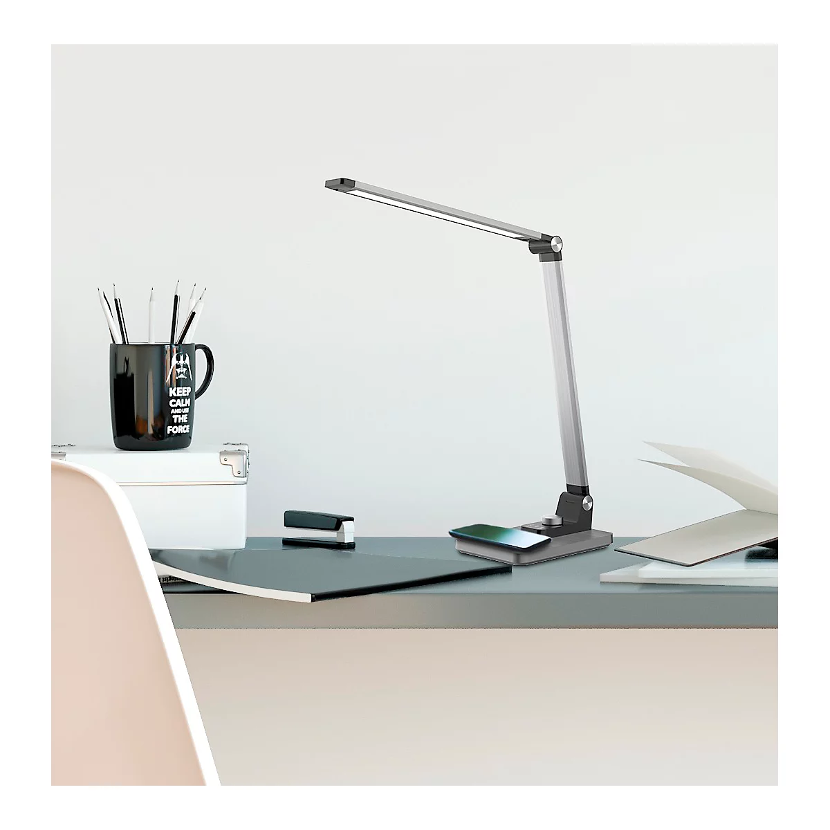 Eine silberne Schreibtischlampe steht auf einem Schreibtisch. Auf dem Schreibtisch sind auch eine Tasse, Hefte und Papier zu sehen.
