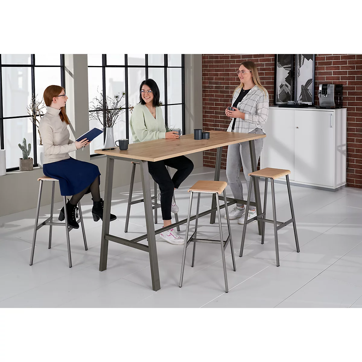 Three women at a high table with stools. One woman is sitting, the others are standing and talking.