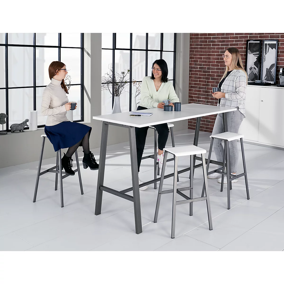 Three women talking at a high table with stools, near a window. Light clothing, gray metal legs.