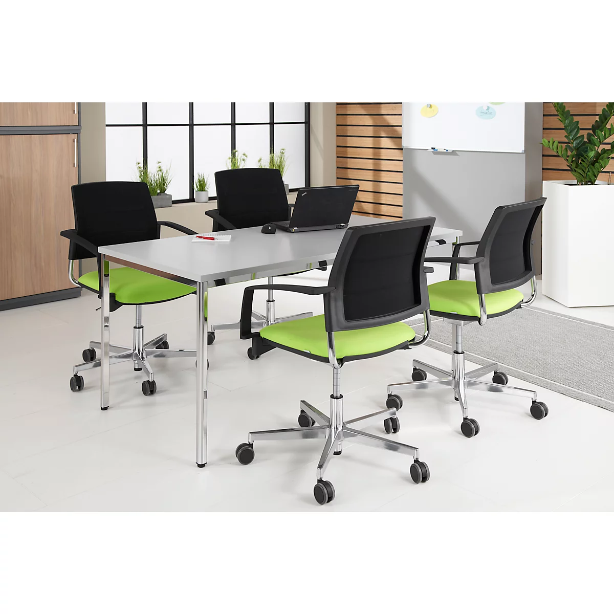 Conference table with 4 chairs. The chairs have a green seat, black backrest and silver frame. There is a laptop on the table.