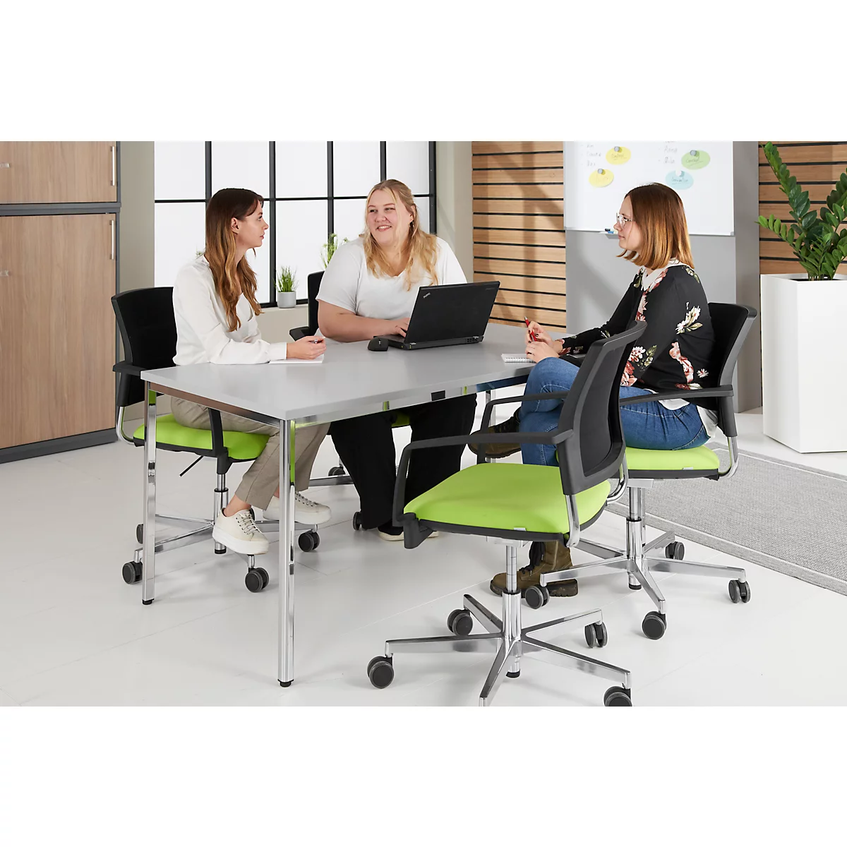 Three women sit around a table. They are talking and working on a laptop.