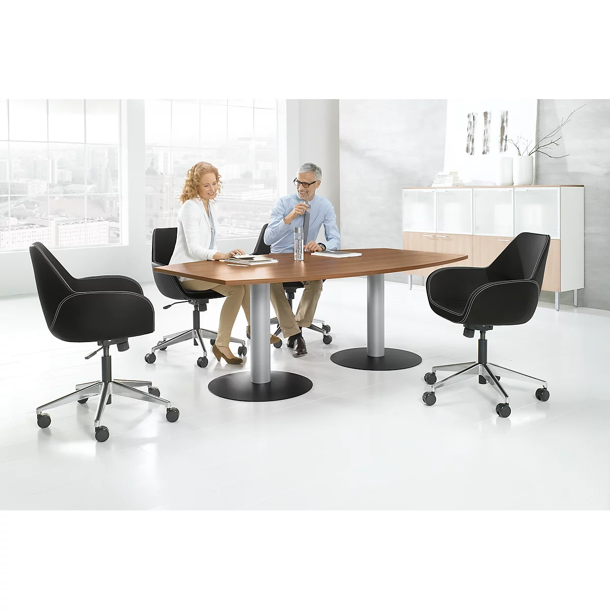 Two people sit at a table surrounded by office chairs. Large windows in the background.