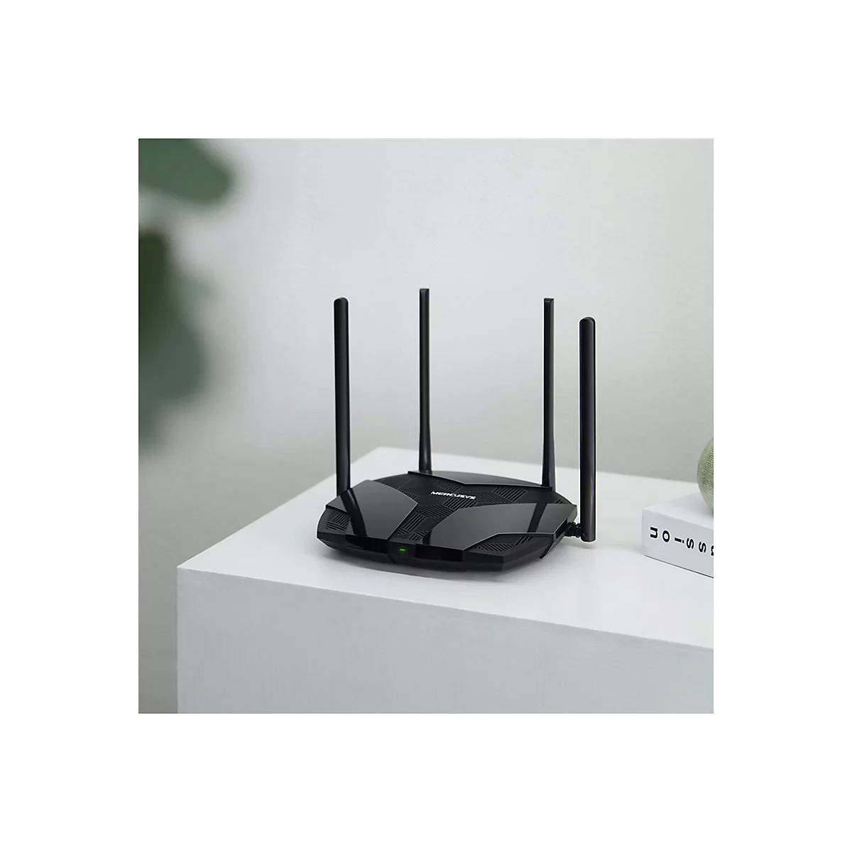Routeur wifi noir avec quatre antennes sur une table blanche. A côté, un livre, en arrière-plan, une plante.