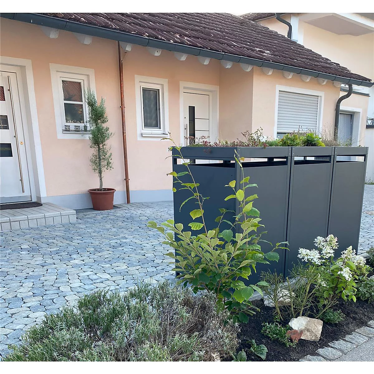 Donkergrijze vuilnisbakkenkast voor een lichtroze huis, omgeven door planten. Kasseien aan de voorkant.
