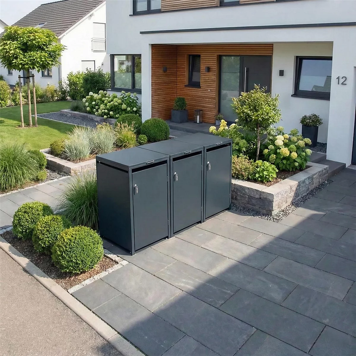 Drie grijze vuilnisbakken voor een modern huis. Geplaveide weg, tuin met struiken en bomen.