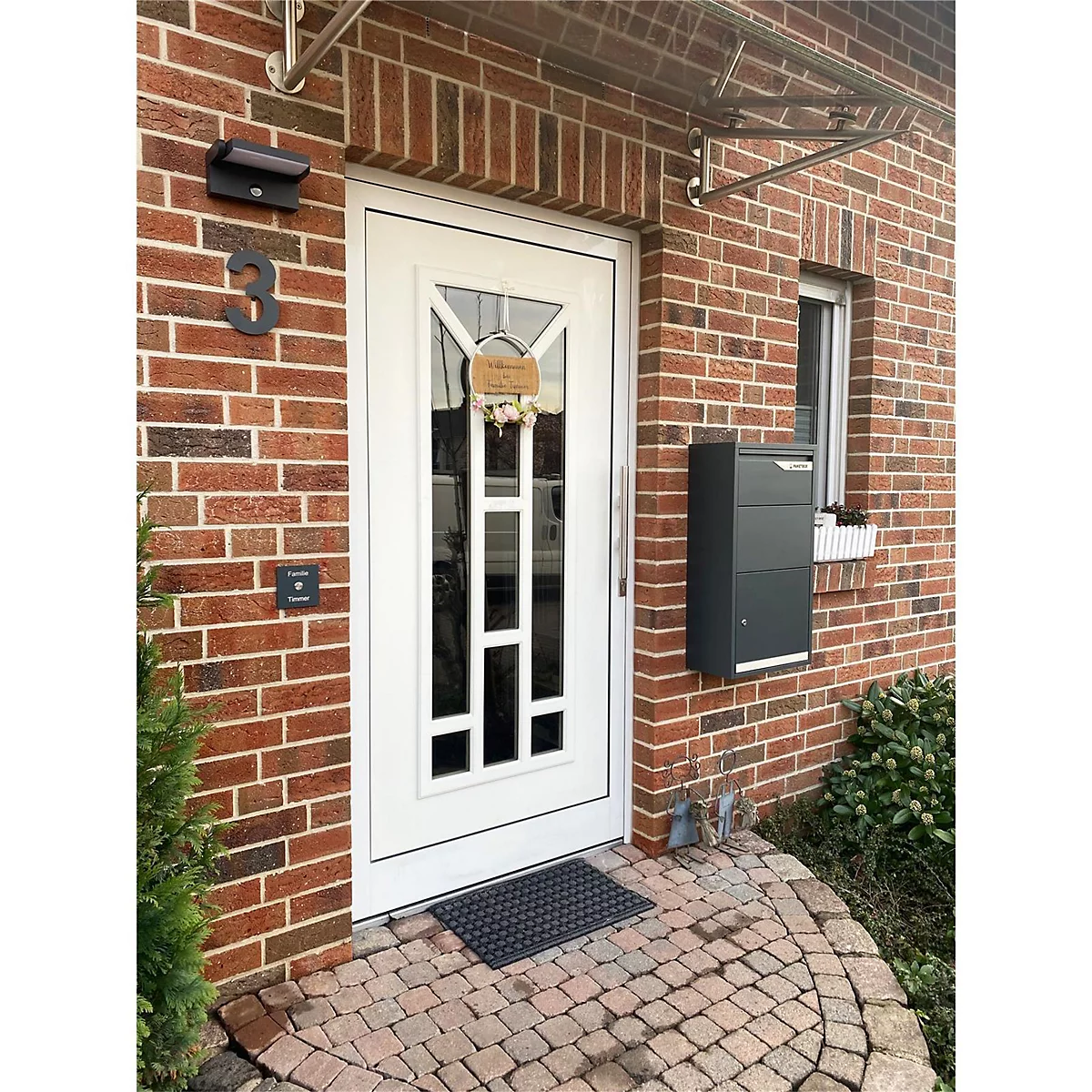 Witte voordeur in bakstenen muur. Een dak boven de deur, daarnaast een brievenbus. Voor de deur een deurmat. Huisnummer 3.