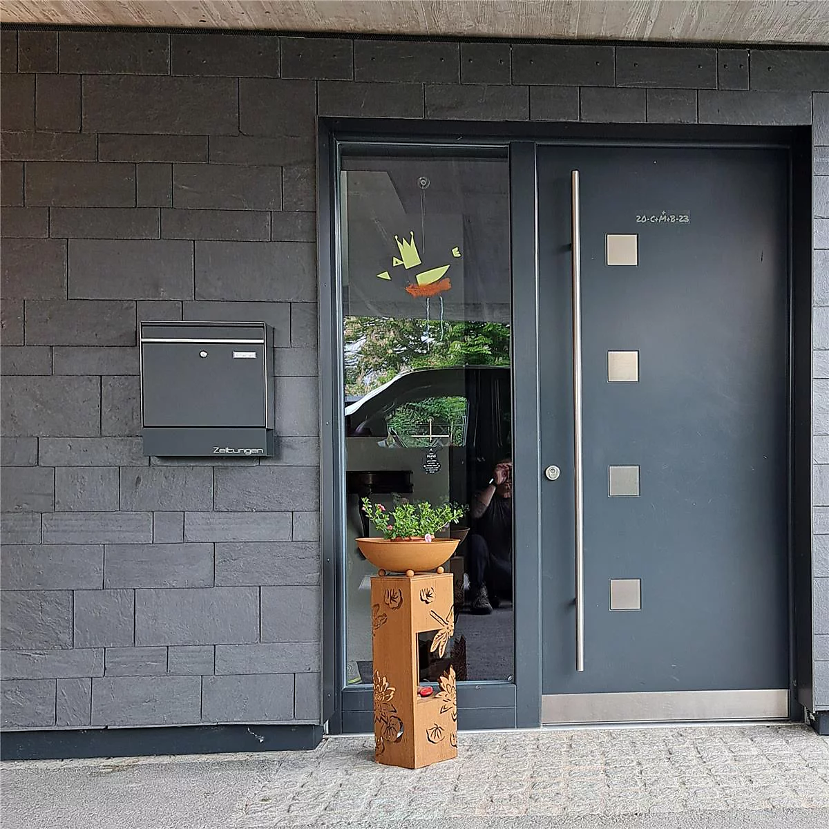 Porte d'entrée gris foncé avec applications en acier inoxydable et boîte aux lettres sur un mur de pierre. Devant la porte se trouve une colonne végétale.