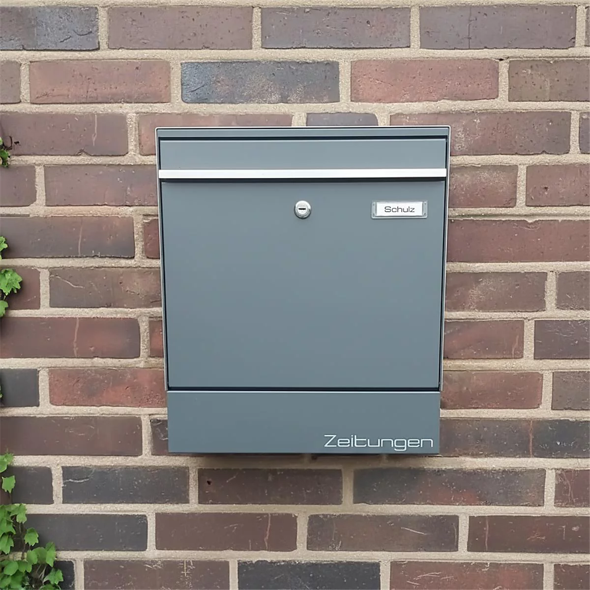 Boîte aux lettres grise sur un mur de briques, avec l'inscription "Zeitungen".