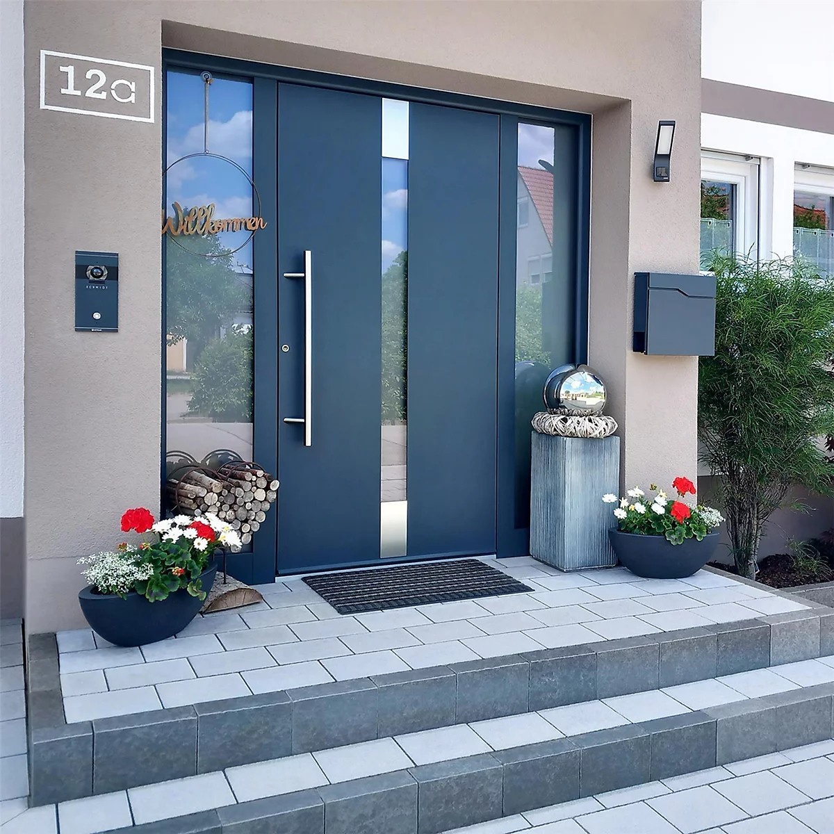 Porte d'entrée bleu foncé avec verre, marches en pierre grise, pots de fleurs, boîte aux lettres et numéro de maison 12a. Scène accueillante.