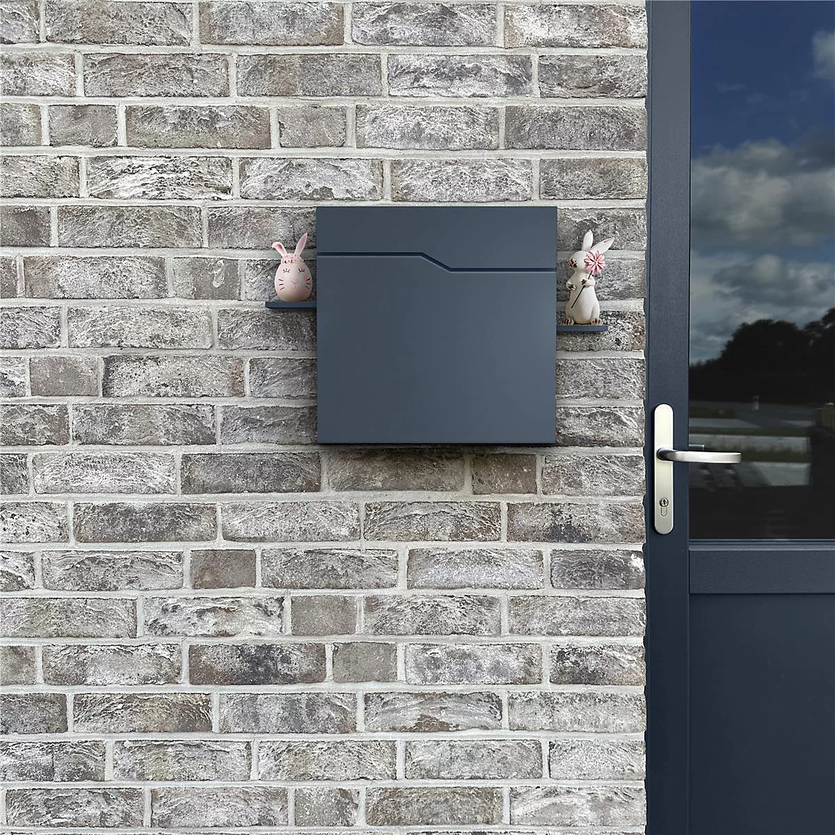 Boîte aux lettres gris foncé sur mur de briques. Deux figurines de lapin la décorent.
