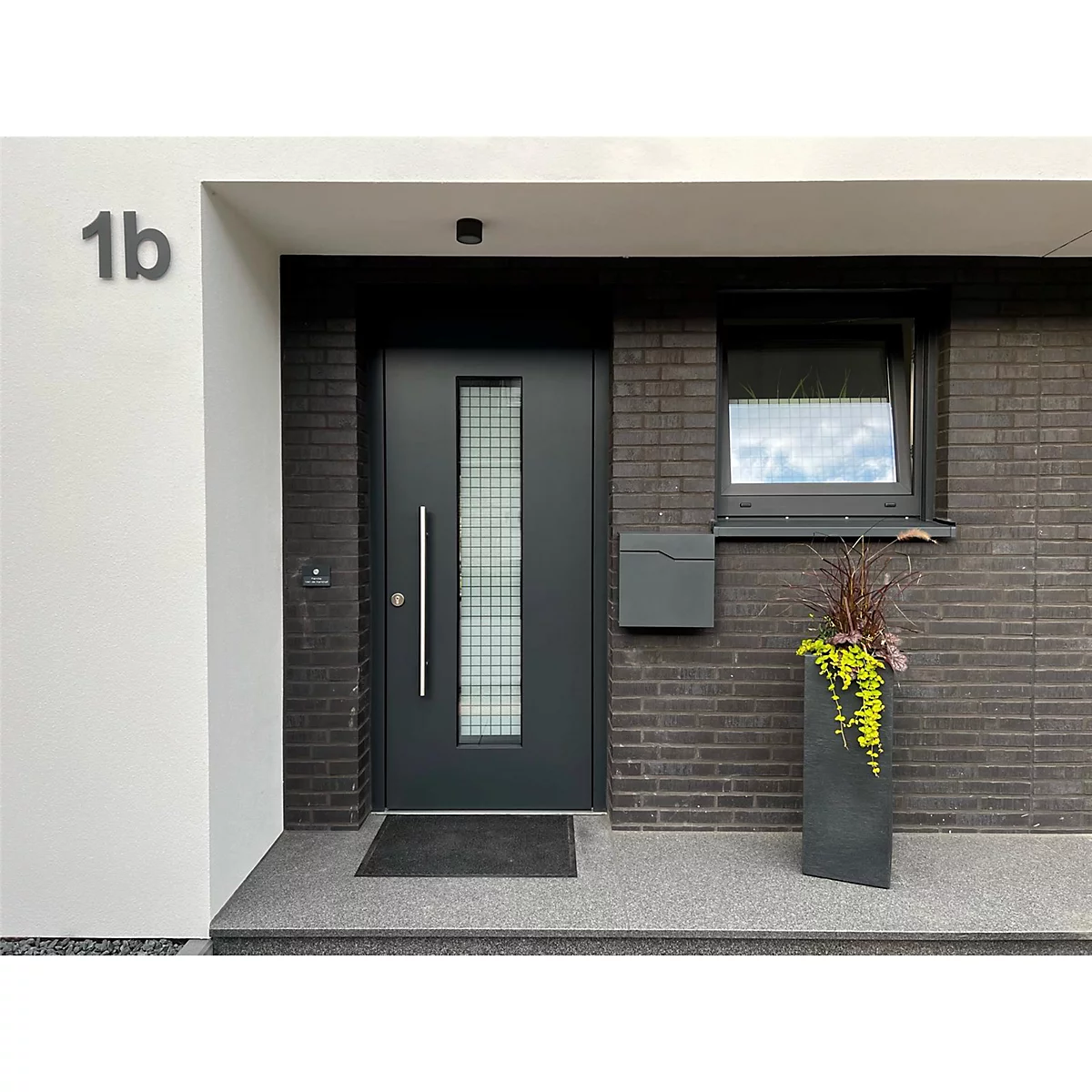Porte d'entrée sombre avec section vitrée et numéro de maison 1b, entourée de briques sombres et de plantes en pot.