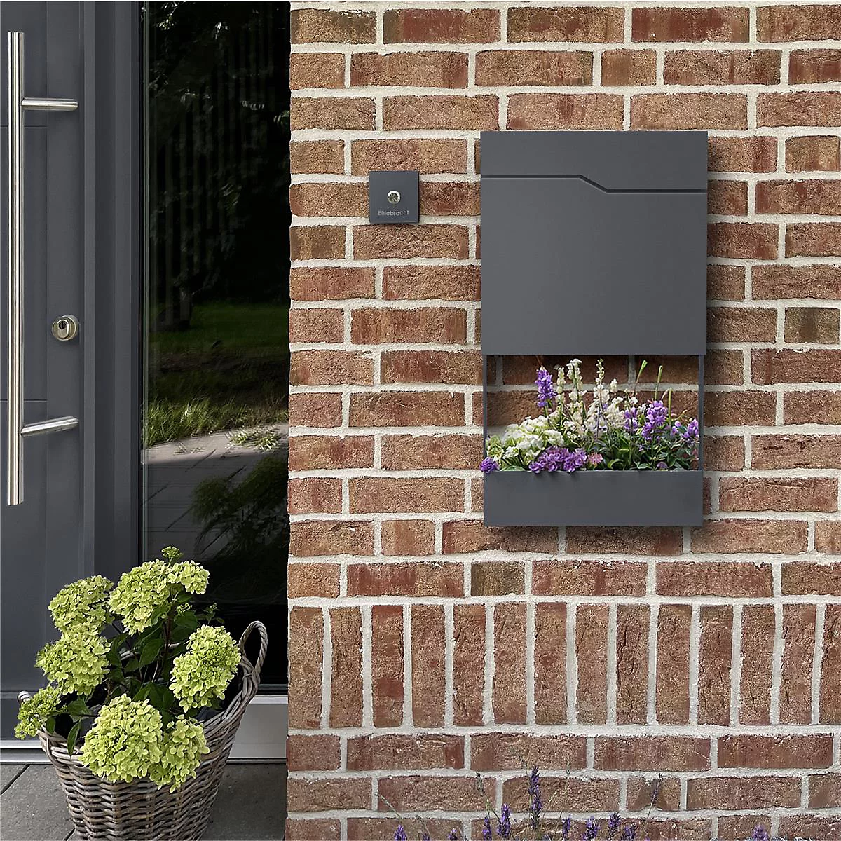 Boîte aux lettres murale gris foncé avec jardinière sur mur de briques. A côté, porte d'entrée et panier avec fleurs. Sonnette au-dessus de la boîte.