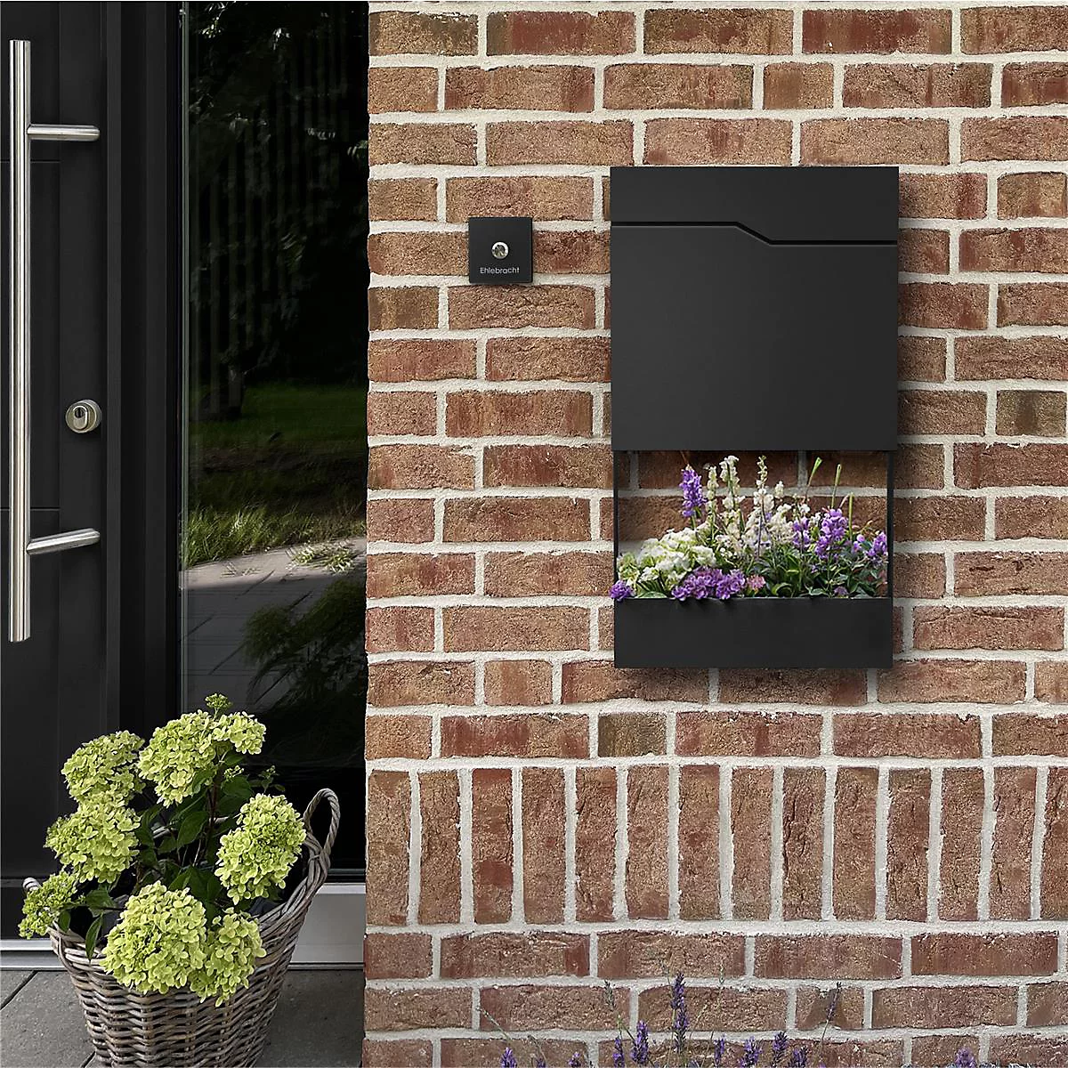 Boîte aux lettres noire avec jardinière intégrée sur un mur de briques, à côté d'une porte et d'un panier de fleurs.