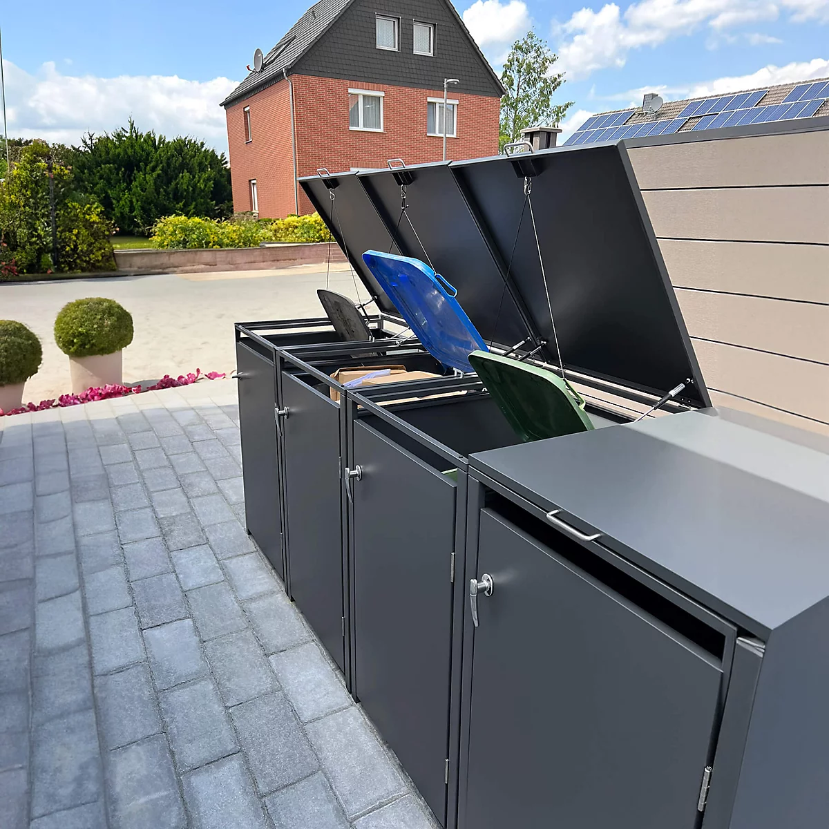 Enclos à poubelles gris avec couvercles ouverts. Poubelles bleues, vertes et noires visibles. Maison en arrière-plan.
