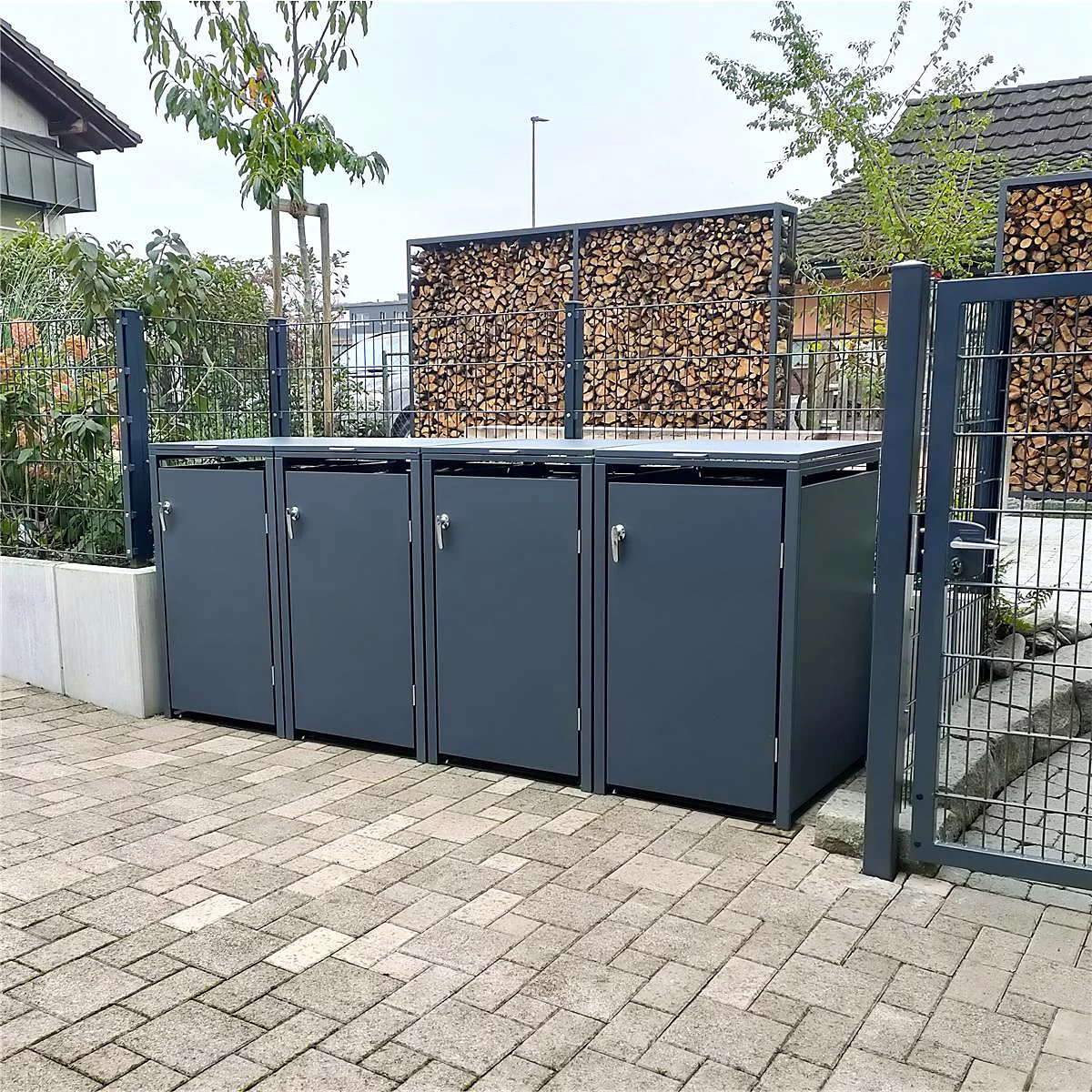 Des bacs à ordures gris sur des pavés, avec un mur en bois et une clôture en arrière-plan.
