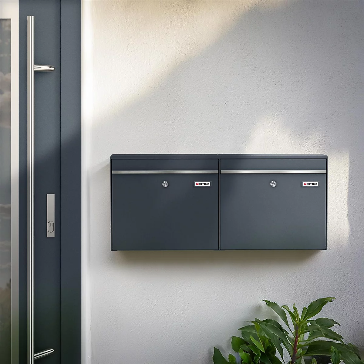 Dunkelgraue Doppelbriefkasten an weißer Wand. Daneben eine Tür. Unten rechts grünes Blattwerk.