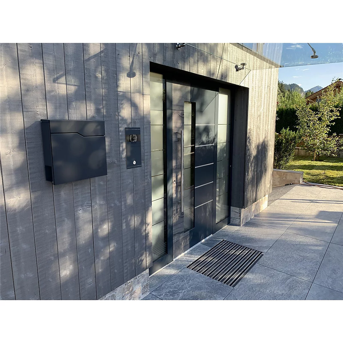 Moderne Haustür mit Klingel und Briefkasten. Der Eingangsbereich ist mit Holz verkleidet. Im Hintergrund sind Berge zu sehen.
