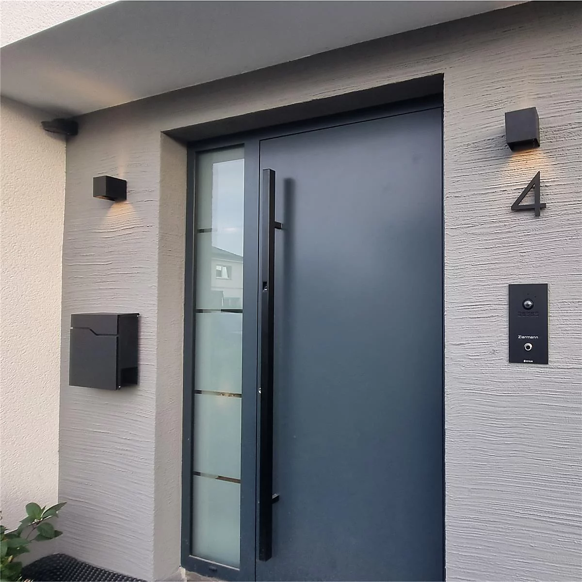 Graue Haustür mit Griff, daneben ein Fenster mit schwarzem Rahmen. An der Wand daneben: Klingel, Hausnummer 4 und Briefkasten.