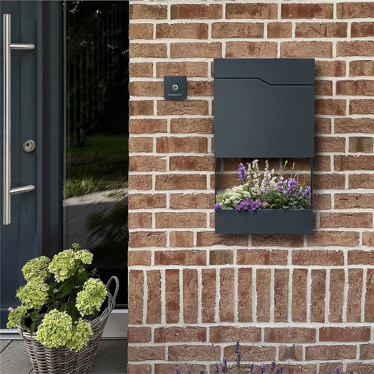 Dunkelgraue Briefkasten mit integriertem Blumenkasten an einer Backsteinmauer. Daneben eine Tür und ein Korb mit Blumen.