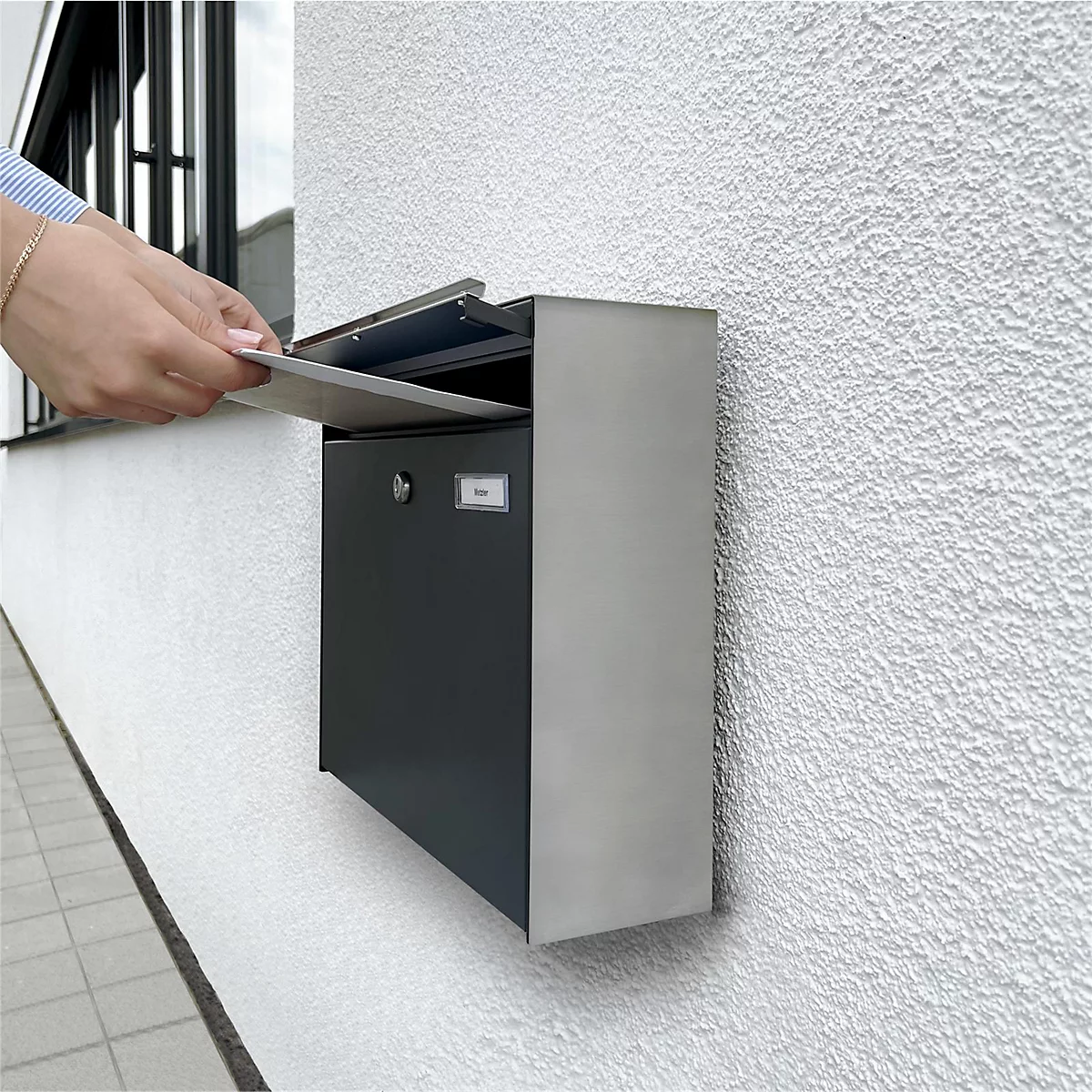 Een hand steekt een brief in een moderne brievenbus aan een huis. De doos is grijs en zilver.