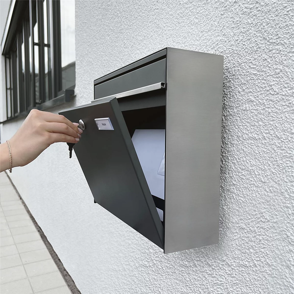 Grijze aan de muur gemonteerde brievenbus met open klep, een arm met sleutel die hem opent. De brievenbus hangt aan een witte muur.