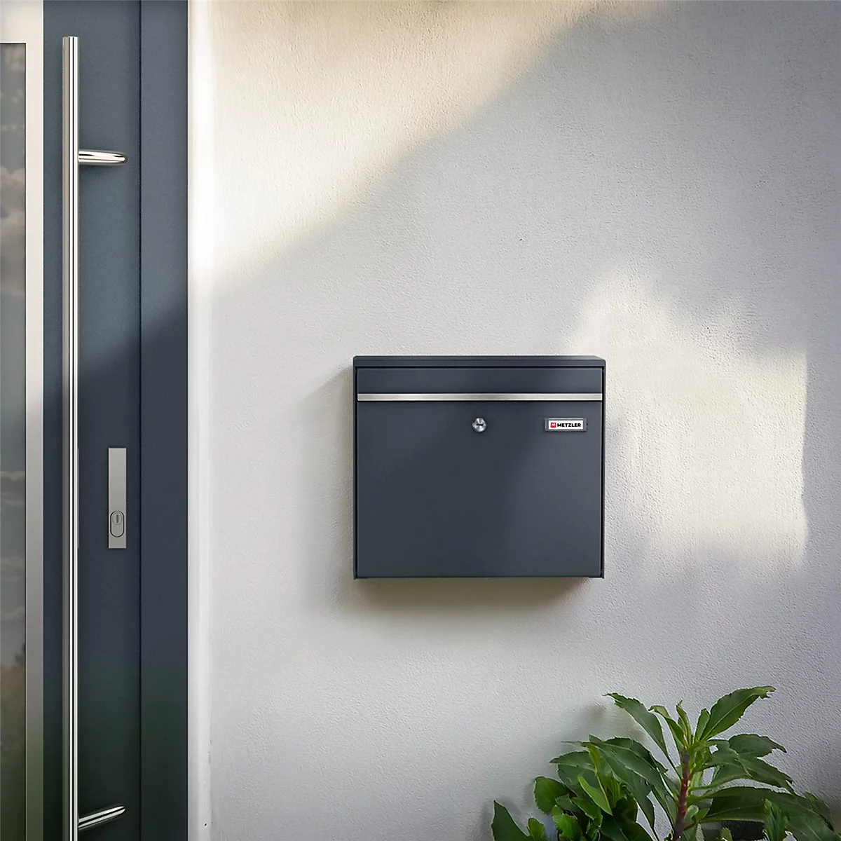 Donkergrijze brievenbus aan een witte muur, groene planten eronder. Daarnaast een grijze deur.
