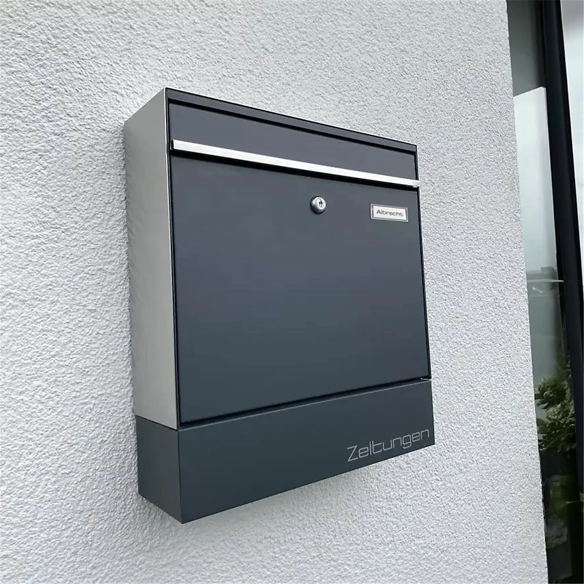 Donkergrijze brievenbus aan huiswand. Zilveren streep en randen. Opschrift: Zeitungen.