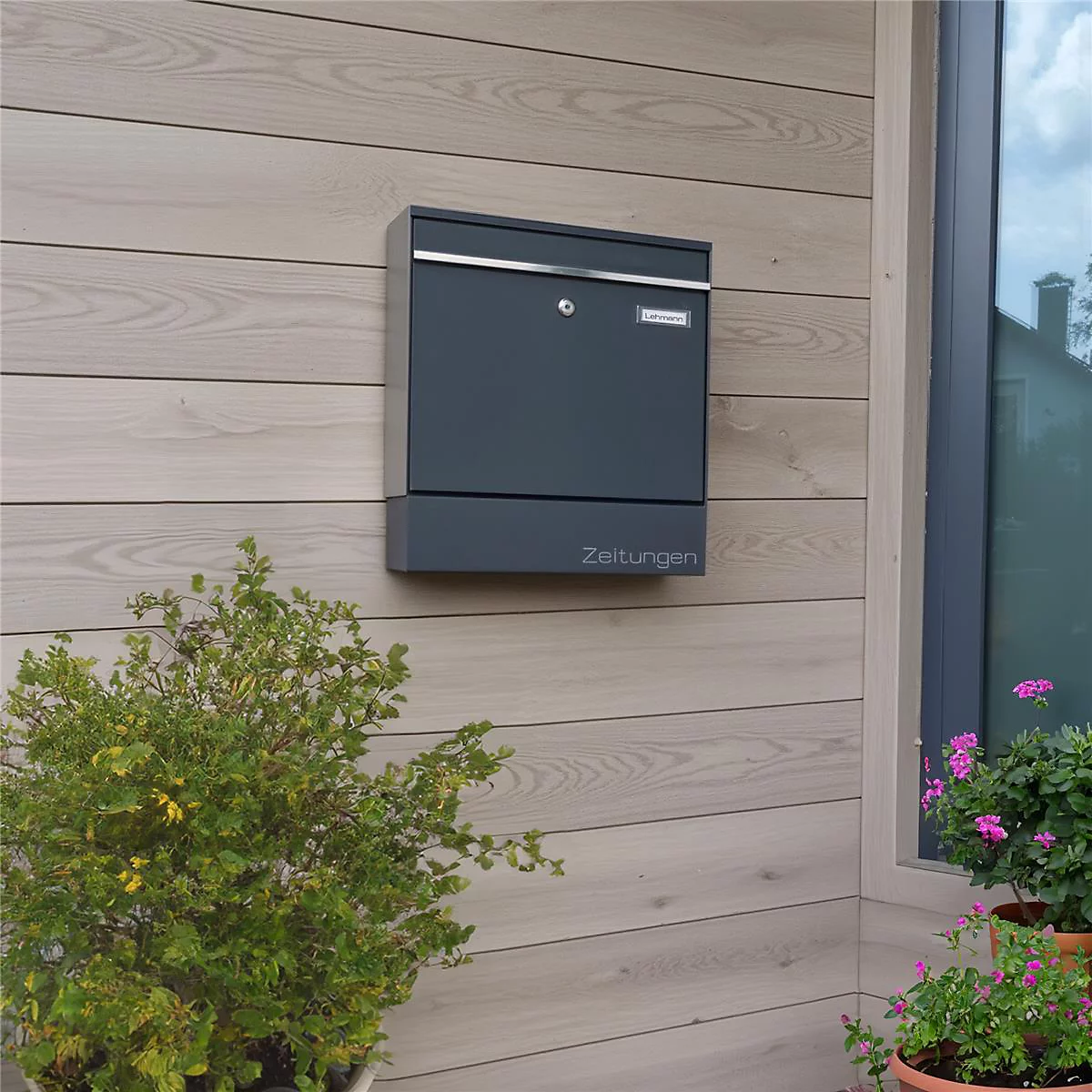 Donkergrijze brievenbus op houten gevel. Inscriptie "Kranten". Rechts een raam met uitzicht op het groen.