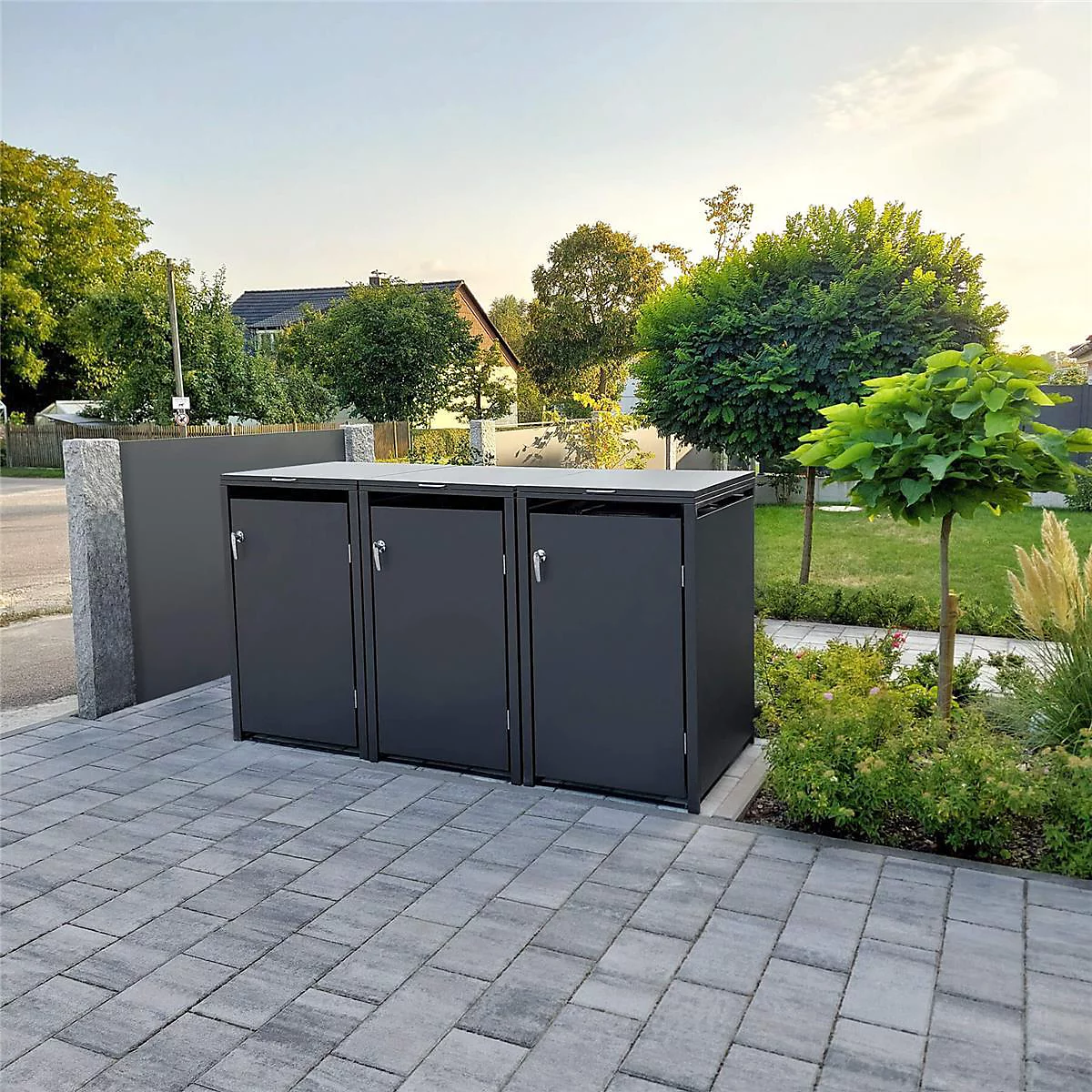 Trois boîtes noires pour poubelles sur des pavés, entourées d'espaces verts et d'arbres.