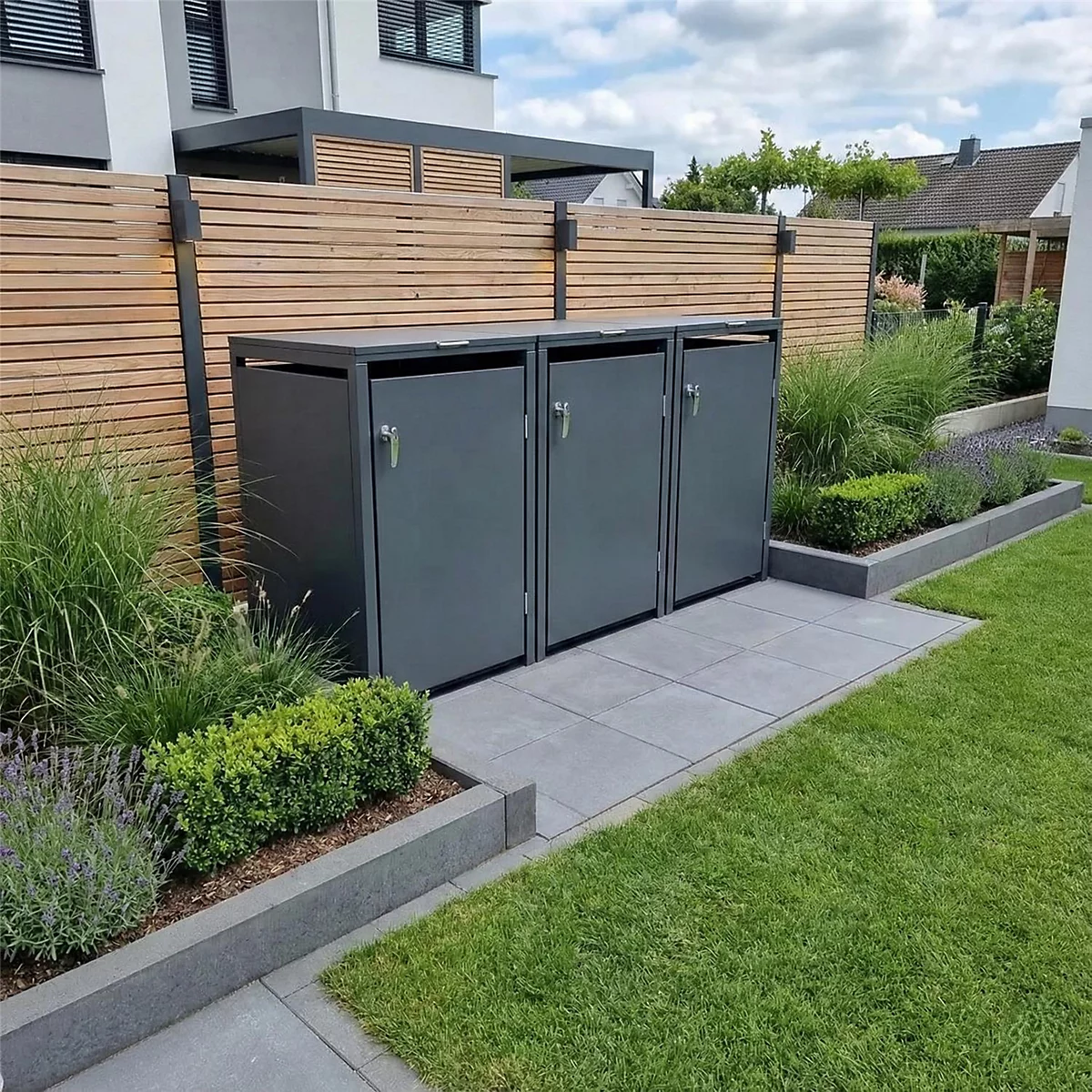 Trois poubelles grises sont dans un jardin devant une clôture en bois, entourées de plantes et de pelouse.