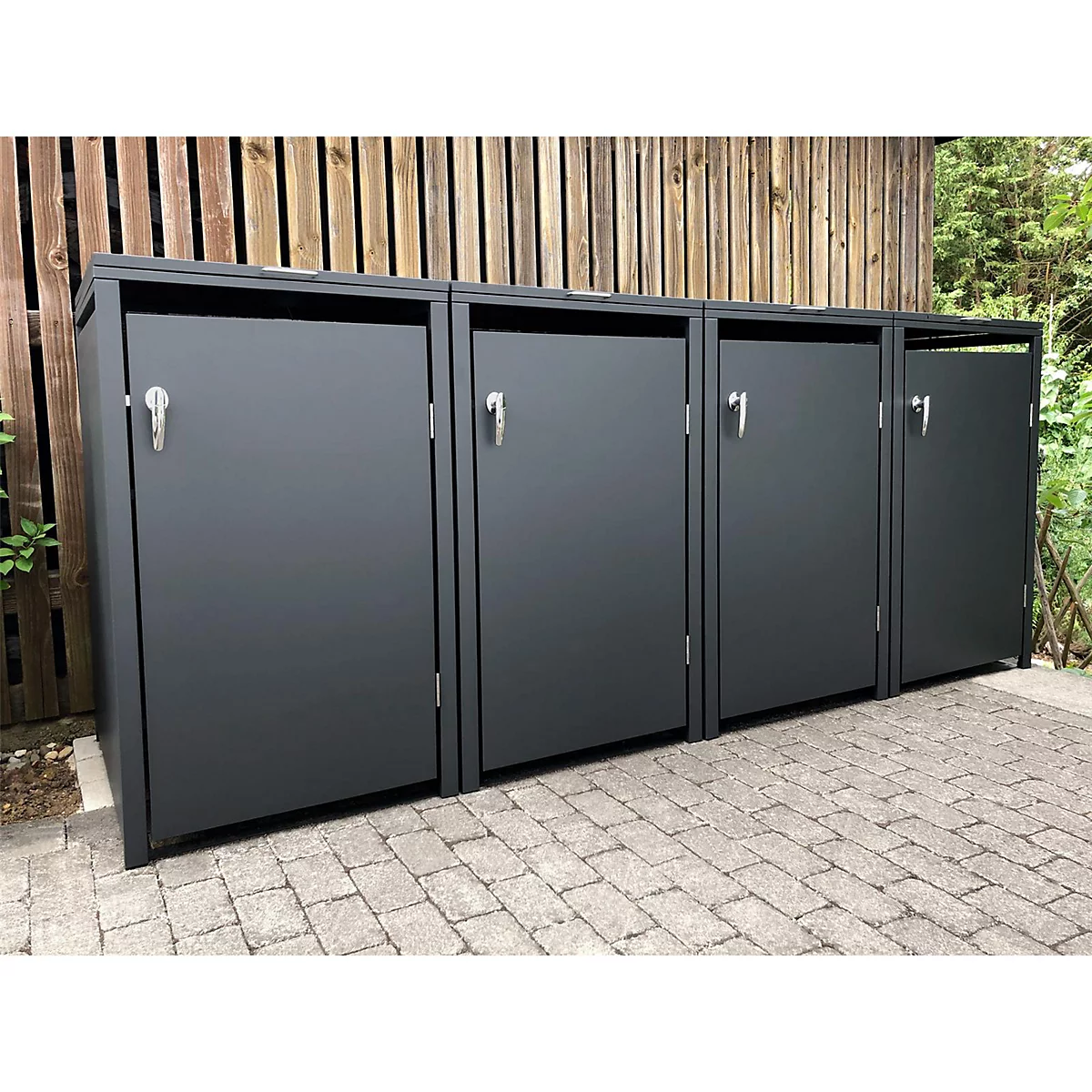 Poubelles grises sur des pavés, devant un mur en bois.