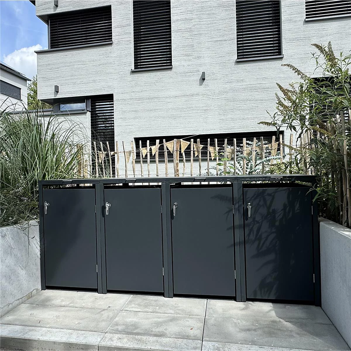 Poubelle sombre devant une maison claire, entourée de plantes. Quatre portes avec poignées.