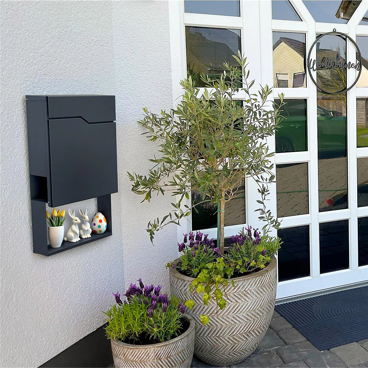 Zwarte brievenbus met plank, paasversiering. Daarnaast een olijfboom in decoratieve potten. Welkomstbord op glazen deur.