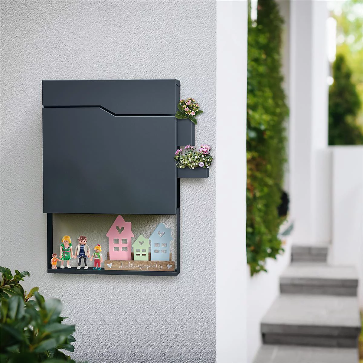 Donkergrijze brievenbus met decoratieve bloembakken. Figuren van familie en huizen eronder. Sfeervol!