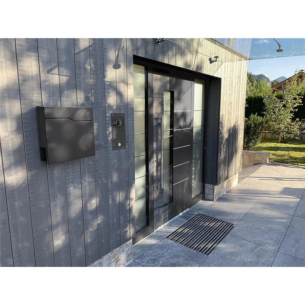 Moderne voordeur met brievenbus en deurbel. De deur is gemaakt van glas en zwart metaal. Grijze houten bekleding.