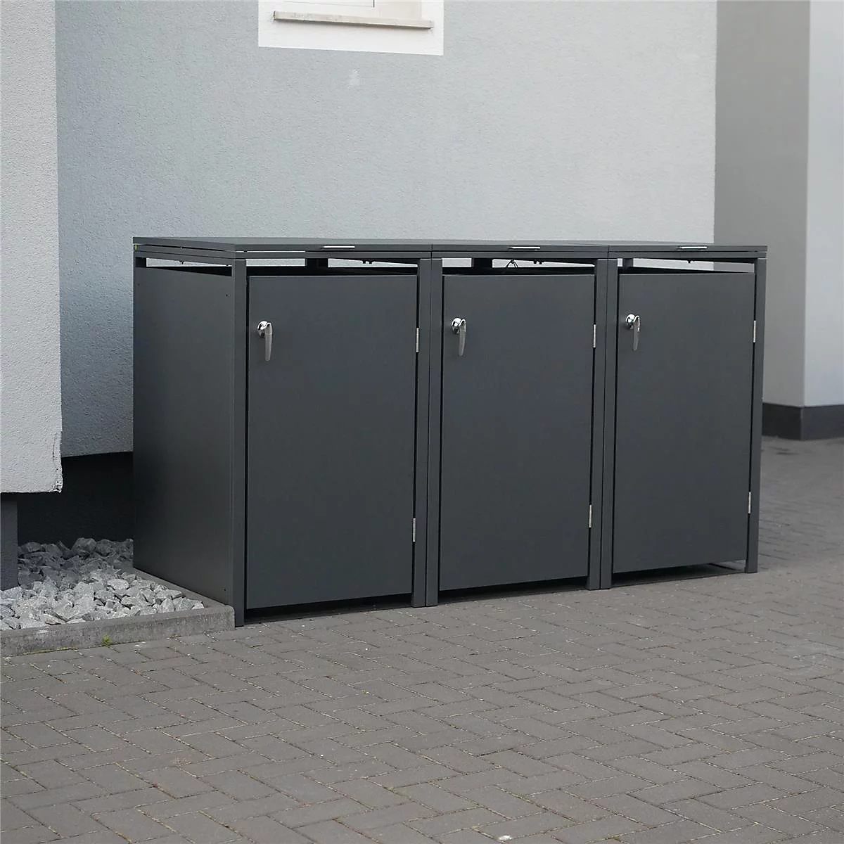 Drei graue Mülltonnenboxen mit Türen und Griffen stehen vor einer Wand.