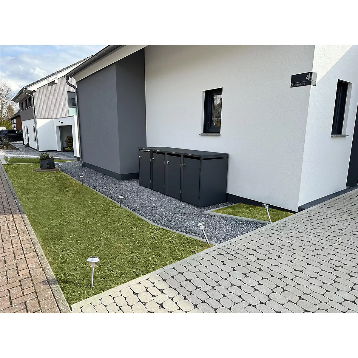 Dunkelgraue Mülltonnenbox vor modernem Haus. Rasen, Kiesweg und Pflastersteine im Vordergrund.