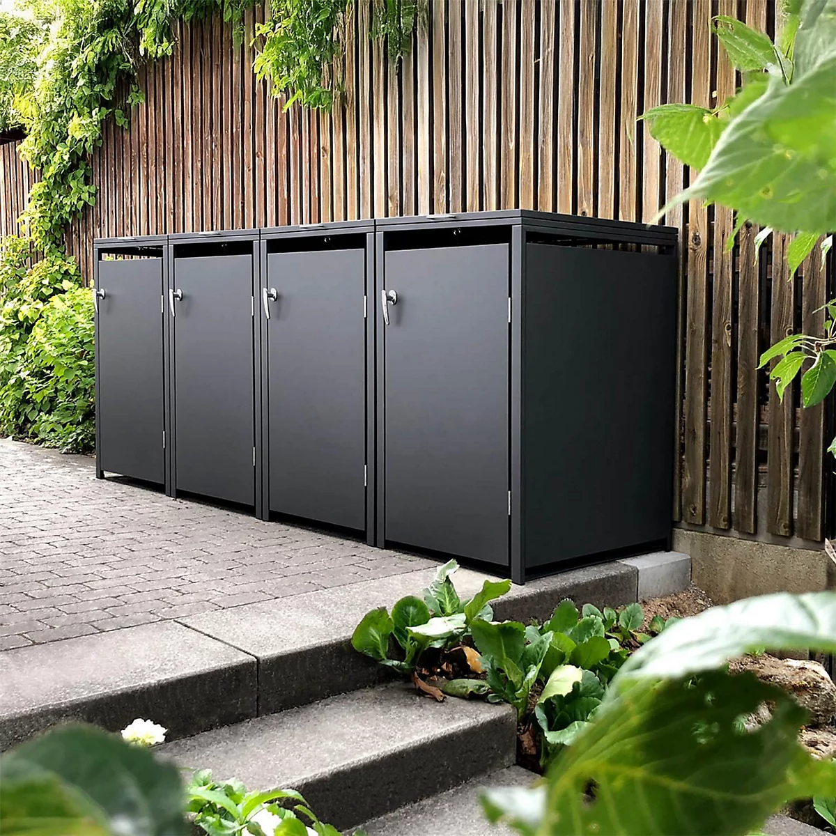 Graue Mülltonnenboxen vor Holzzaun. Grüne Pflanzen umgeben die Boxen.