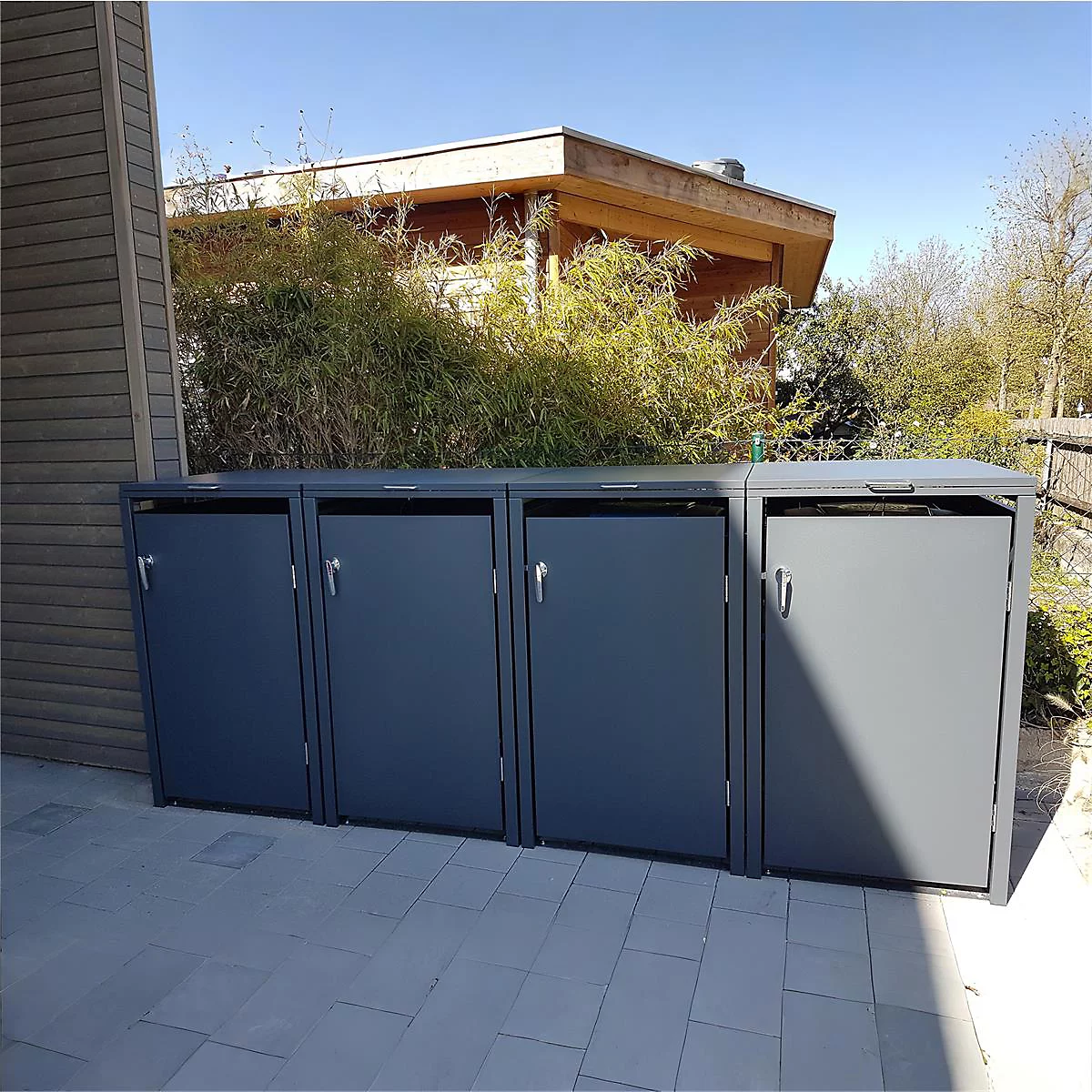 Graue Mülltonnenboxen auf Steinplatten, dahinter Büsche und ein Haus.
