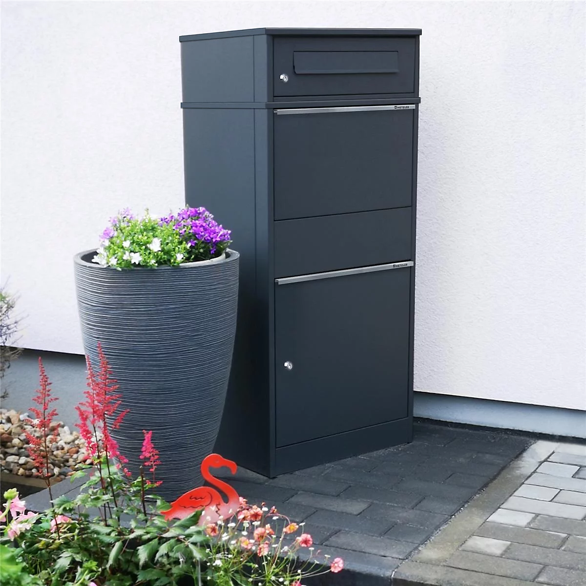Dunkelgrauer Briefkasten mit drei Fächern, neben einer Blumenvase mit bunten Blüten. Am Boden ein roter Flamingo.