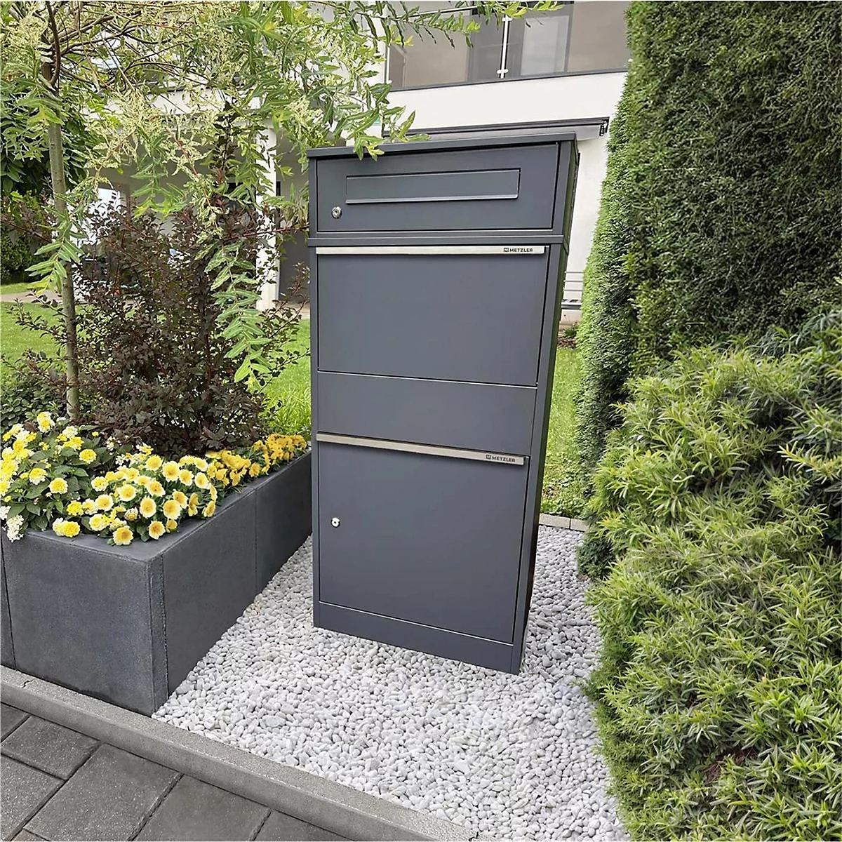 Dunkelgrauer Briefkasten mit drei Fächern, steht auf Kies neben Blumen und Büschen.