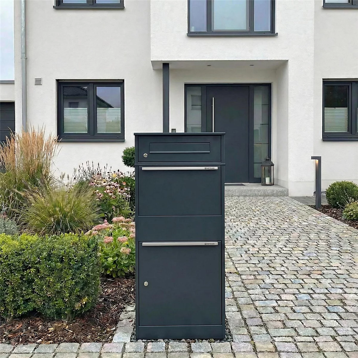 Dunkelgraue Briefkastenanlage mit zwei Fächern und einem separaten Fach darüber. Sie steht vor einem Haus mit Garten.